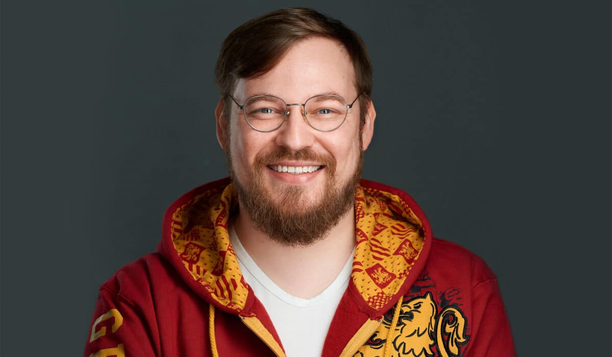 A man with a beard and glasses, wearing a red hoodie with a detailed yellow pattern, smiling against a dark background.