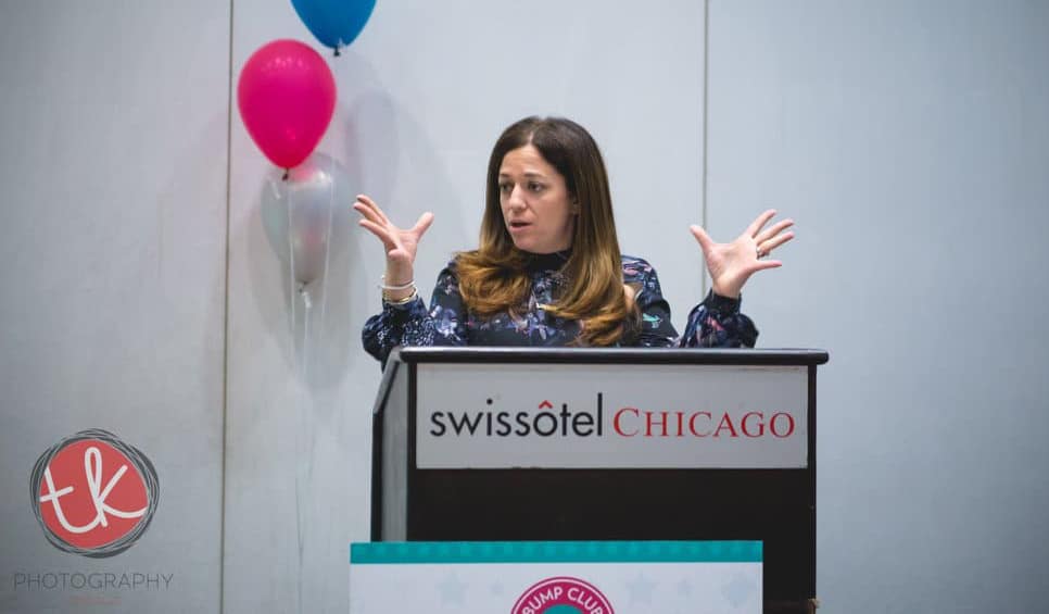 A woman stands at a podium that reads "swissotel Chicago". The woman has her hands in the air, fingers spread out, and maintains a serious look on her face as she delivers a lecture.