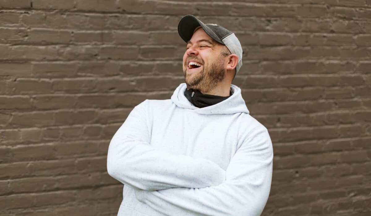 Chris Coyier, in a white sweater and hat, is laughing in front of a brick wall