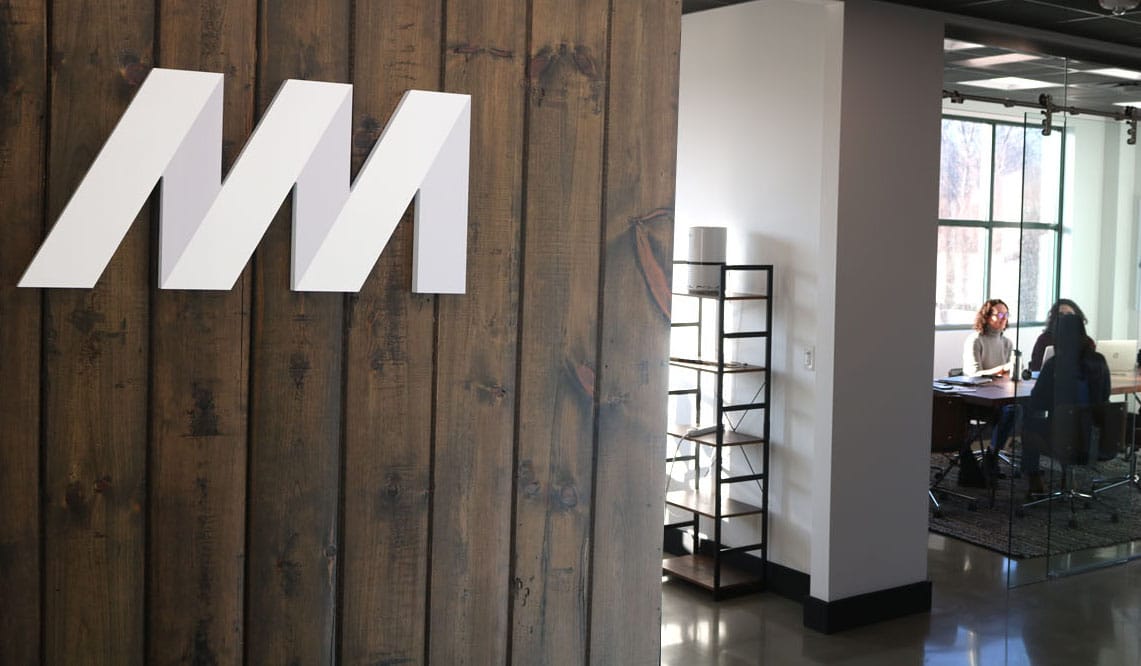 A logo for Newor Media sits on a wooden-paneled wall. To the right in the distance are colleagues sitting at a desk together.