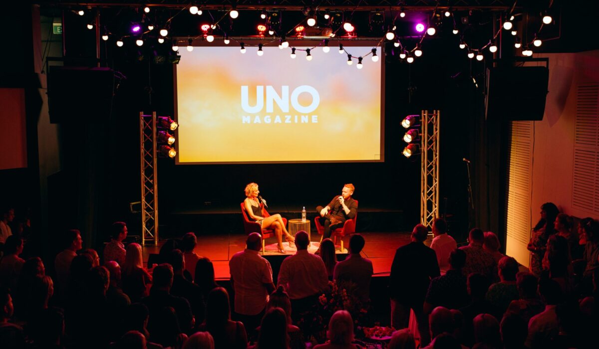 Two people holding microphones sit on a stage adorned with UNO Magazine on a presentation in the background