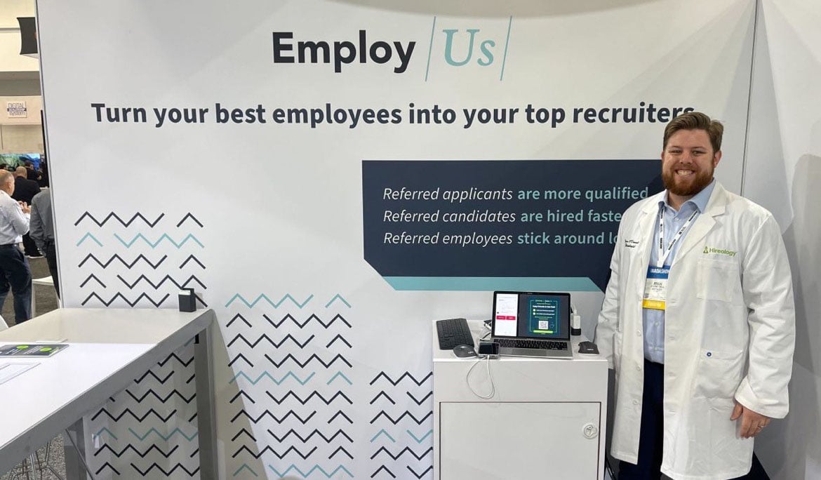 A man stands in a white coat smiling at the camera, in front of a large banner that says EmployUs