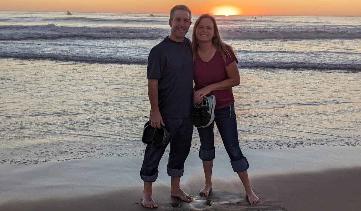 Two people stand barefoot on a beach at sunset smiling at the camera. In the background is an orange sunset reflecting off the waves of a beach.