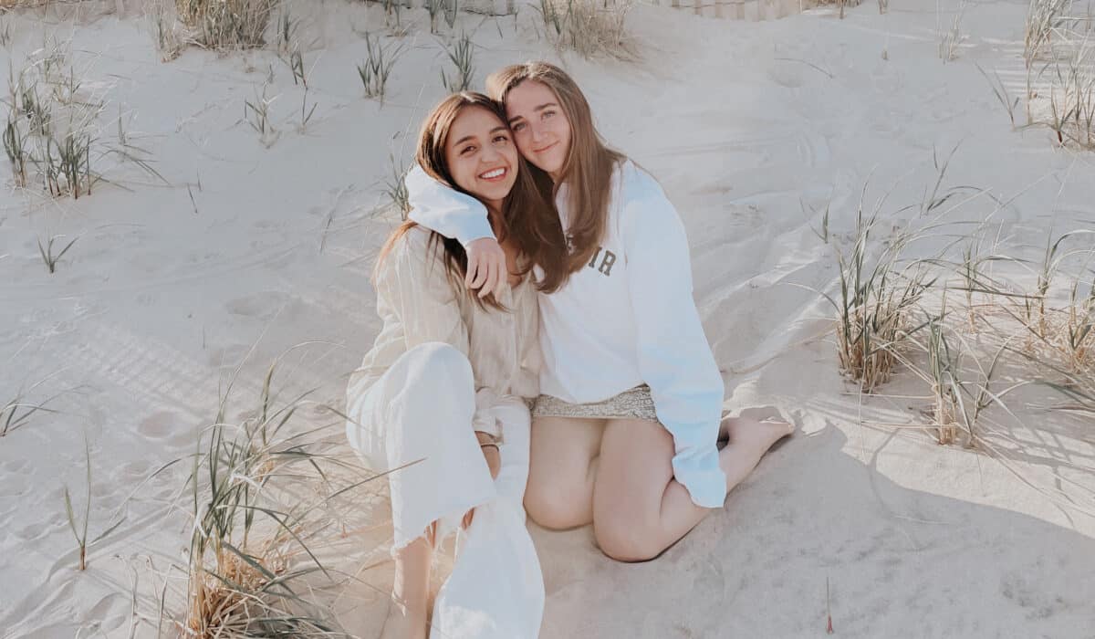 Two females sit side-by-side in the sand, smiling at the camera