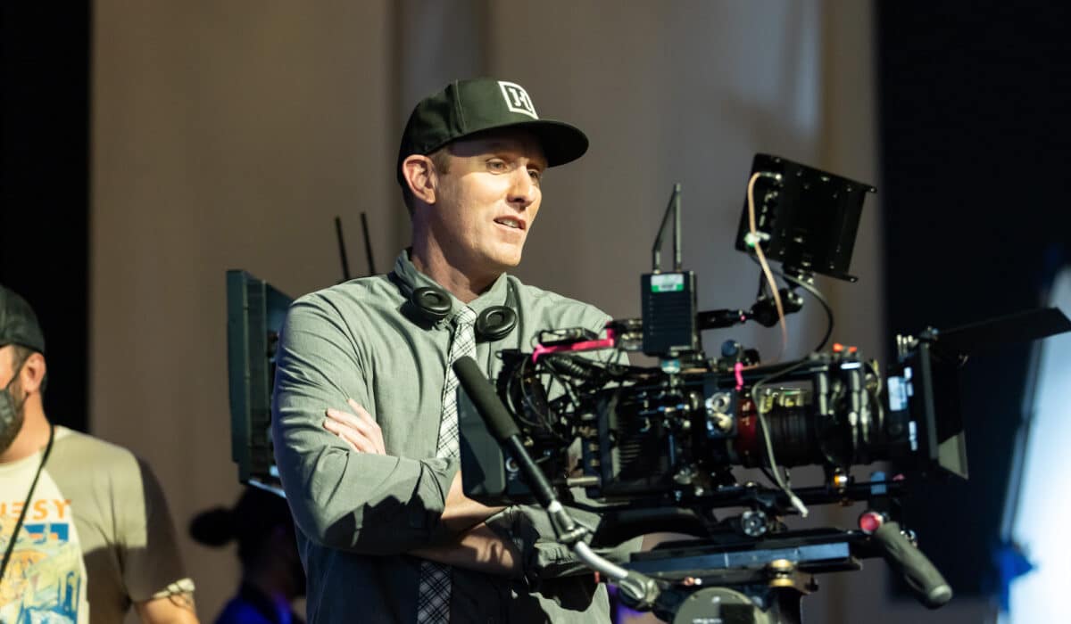 A person in a baseball cap stands in front of a large camcorder on a film set.