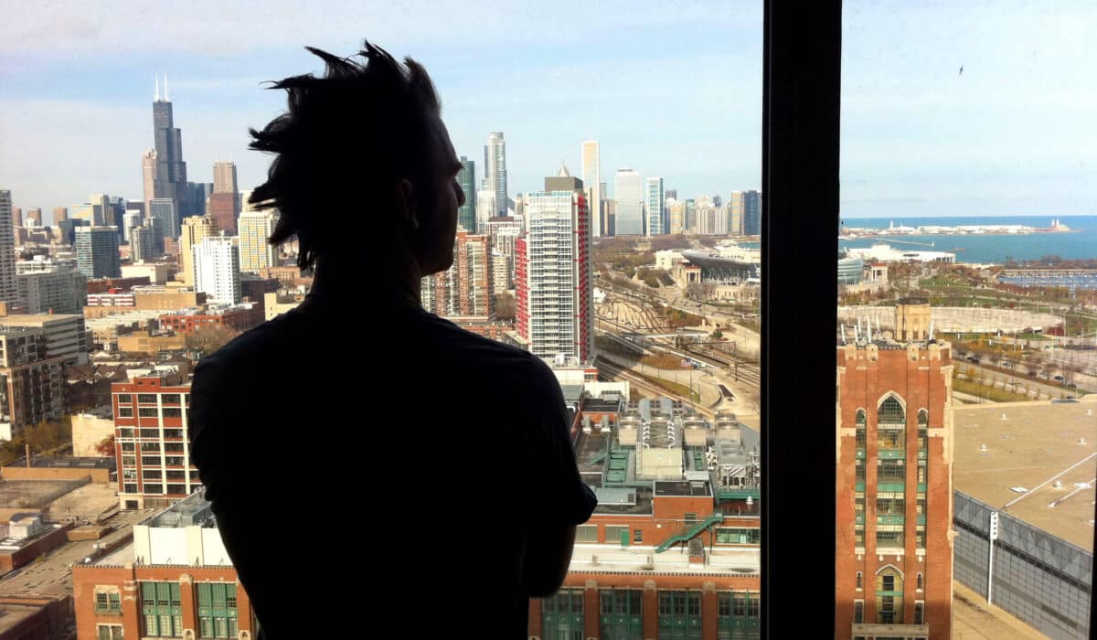In the foreground is the shadow of a person with spiky short hair. In the background is a cityscape with tall buildings and a clear blue sky.