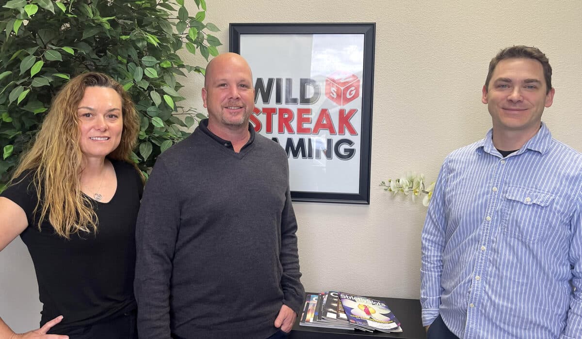 Three business people stand in front of the camera, smiling. In the background is a tall plant, and a framed picture sporting the "Wild Streak Gaming" logo.