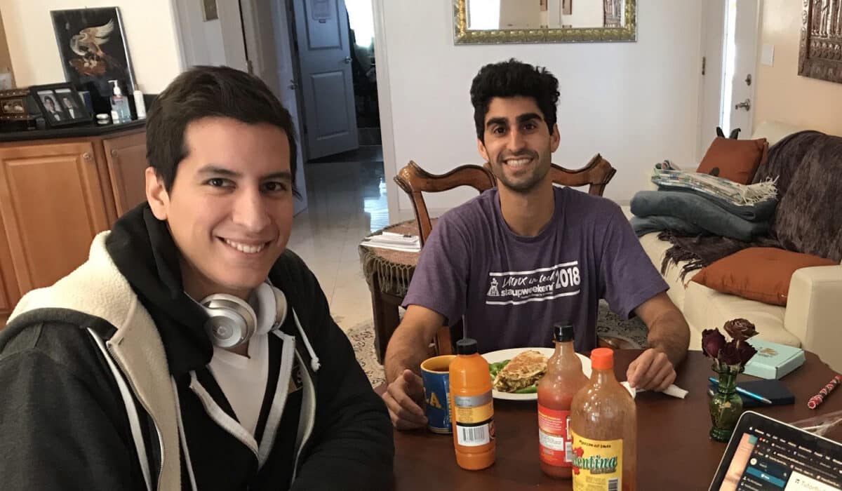 Two men sit at a table with hot sauce, smiling at the camera. The person on the left wears a black hoodie sweater with headphones around his neck. On the right, he wears a purple t-shirt.