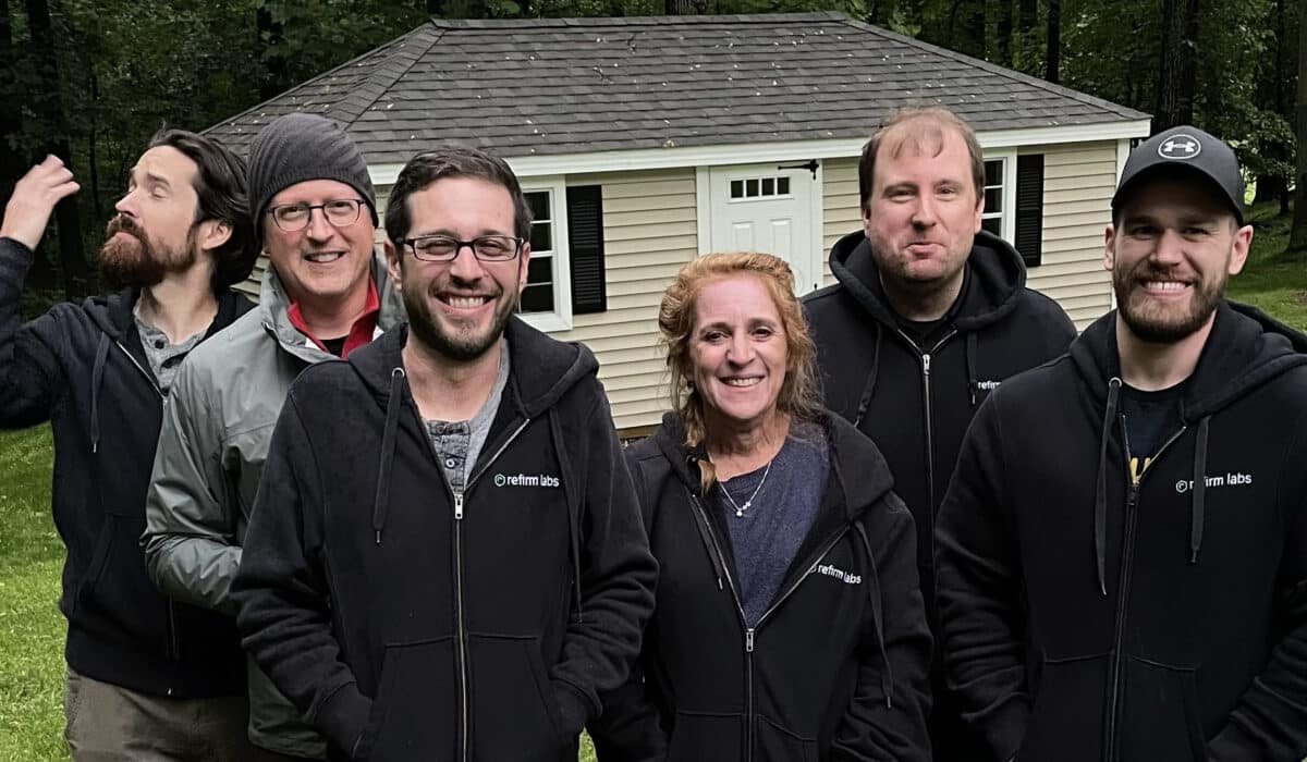 A group of six people stand outside smiling at the camera. Each person wears a black, zipped-up sweatshirt that has the logo ReFirm Labs embroidered on it.