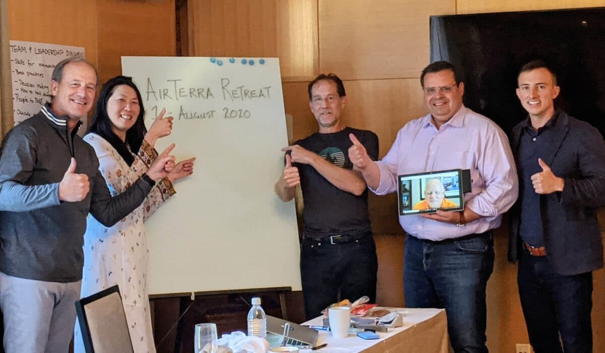 Six businesspeople stand in a line with their thumbs up at the camera. Behind them is a whiteboard with the words "AirTerra Retreat" written down in marker.