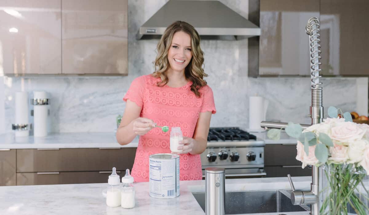 A woman in a pink t-shirt stands in front of a marble kitchen counter scooping formula into a clear bottle. On the counter are two additional bottles of formula and a big tub of formula.