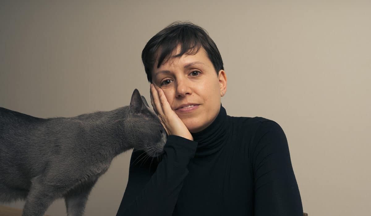 A business person in a black turtleneck is centered with their head resting on their hand. On the left is a grey cat, brushing up again the business person's hand.
