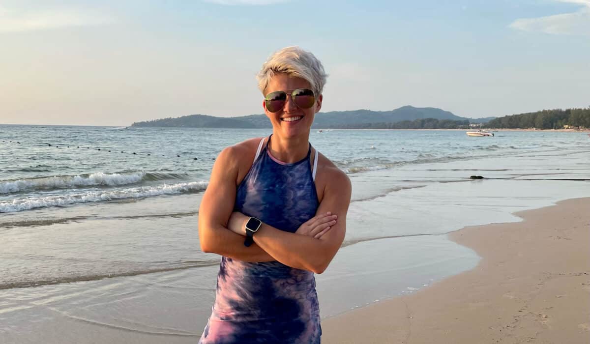 A woman with sunglasses and a purple dress stands with her arms crossed, smiling at the camera. In the background is a beach.