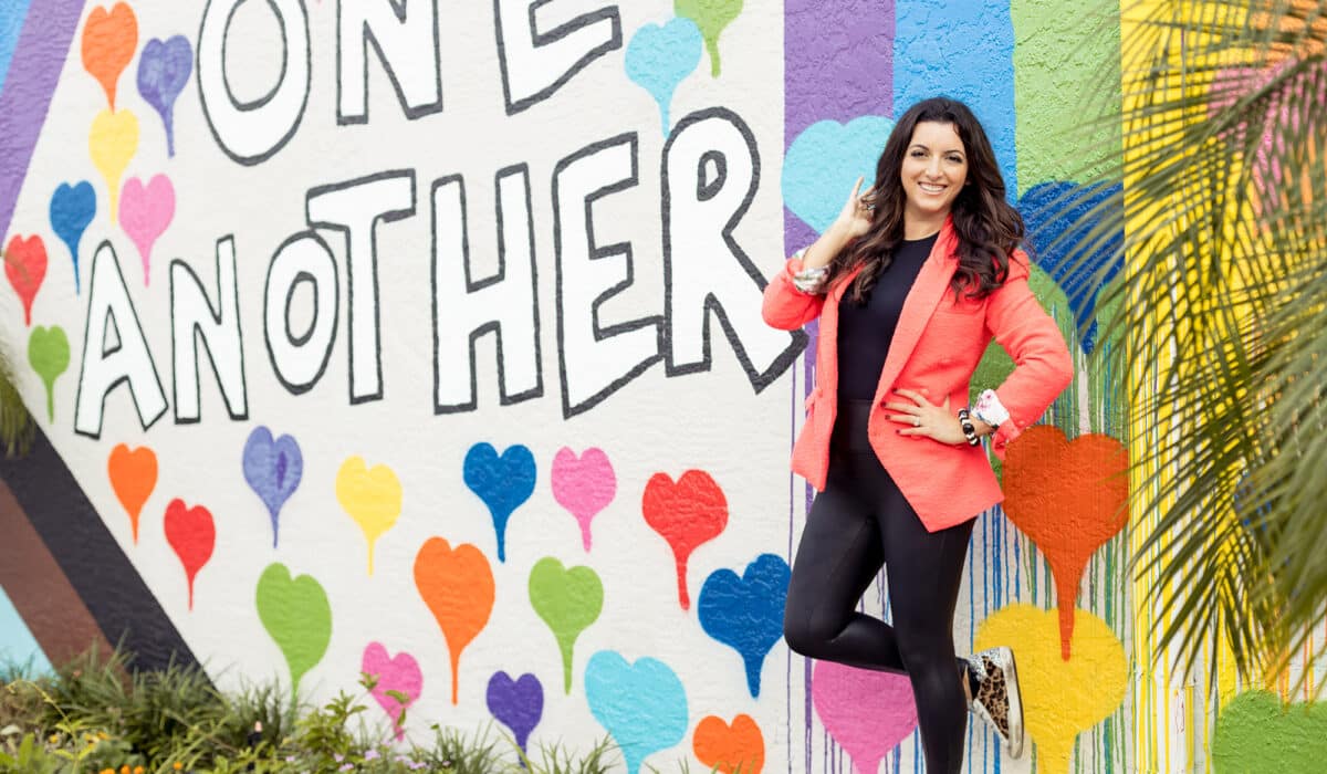 A woman standing in front of a wall with colorful hearts. She is wearing a pink jacket and black tights, and has flowered socks on her feet. There are also words "Love One Another" painted on the wall, as well as blue drops and pink hearts scattered around the surface. The dominant colors in this image are white and pink.