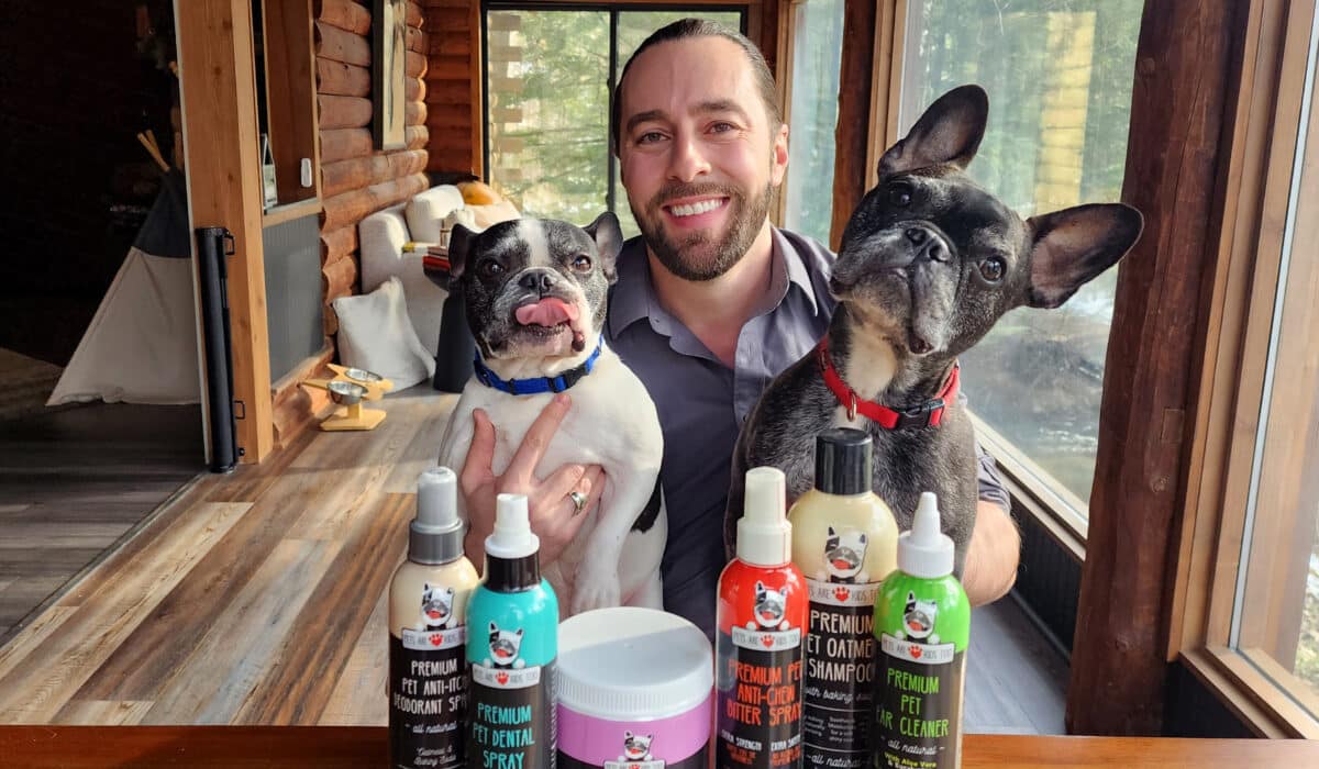 A man smiles at the camera while holding two dogs. In the foreground are a bunch of dog products in tubes and jars.