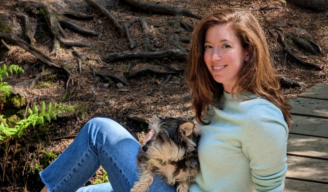 A young woman with long, dark hair is sitting on the ground in a woodsy area with light blue sweater and blue jeans, and she has a bright smile on her face as she looks directly at the camera. In her lap sits a terrier dog.