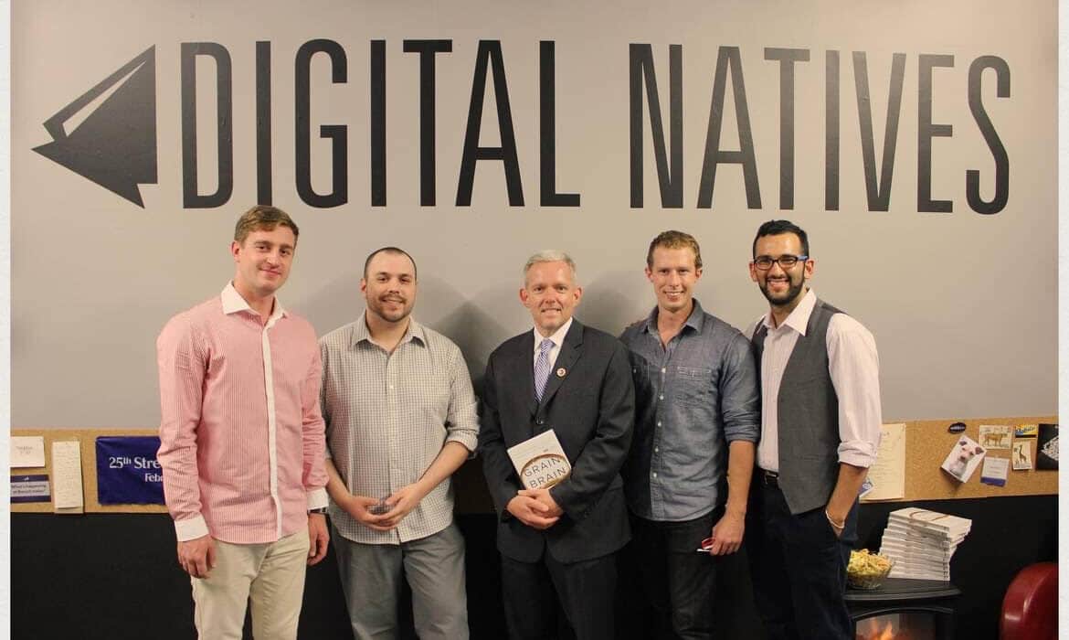 A group of men stand in line in front of a wall with the "Digital Natives" logo embedded on it.