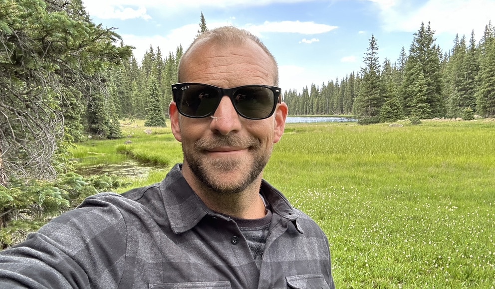 A man is smiling at the camera, wearing sunglasses and a grey checkered shirt, with lush pine trees and a pond in the background.