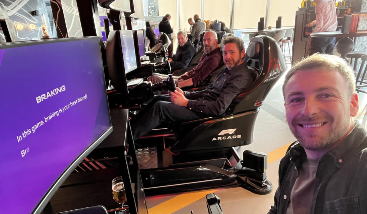 A group of men enjoy a simulated racing experience in an arcade, with one taking a selfie in the foreground and the screen displaying a message about braking in the game.