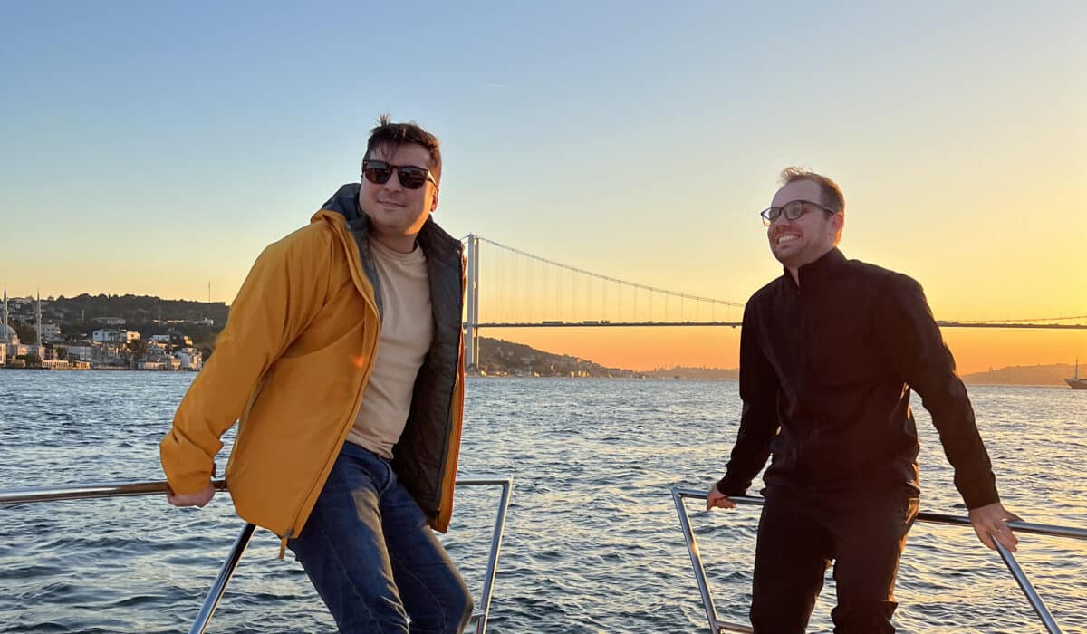 Two men are enjoying a boat ride with one wearing sunglasses and a yellow jacket, and the other smiling in a black turtleneck, with a suspension bridge and sunset in the background.