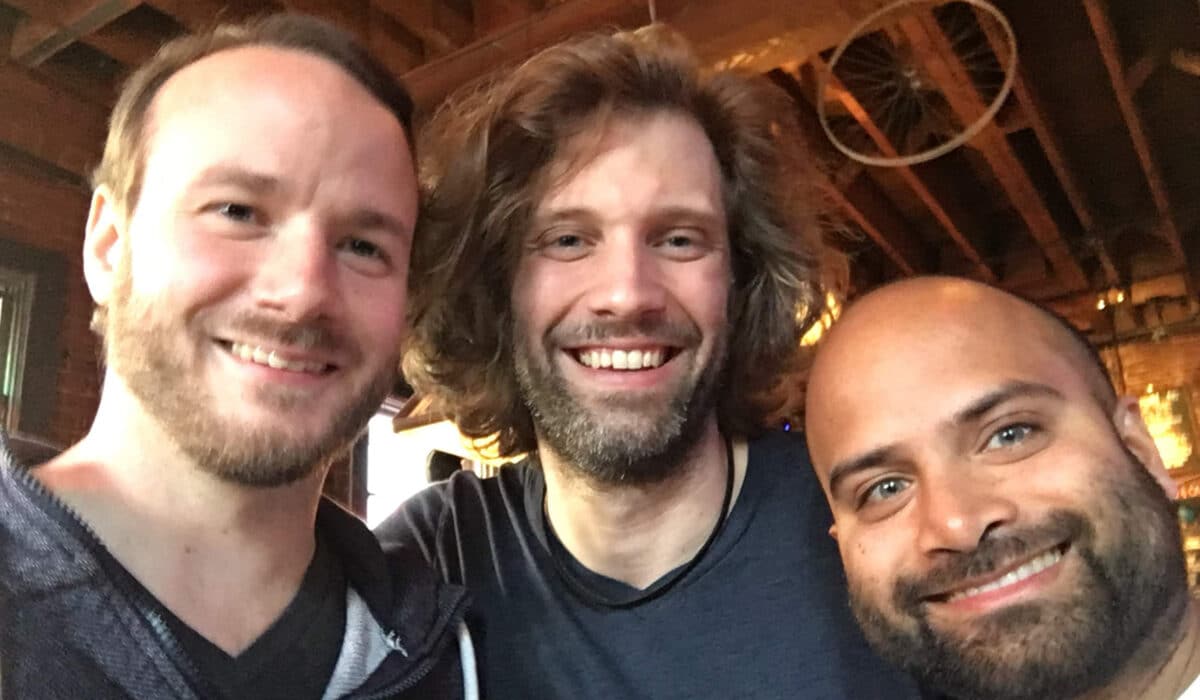 Three smiling men posing closely together for a selfie, with one sporting a beard and black V-neck, the middle with long wavy hair and a tank top, and the third with a shaved head, in a room with rustic decor.