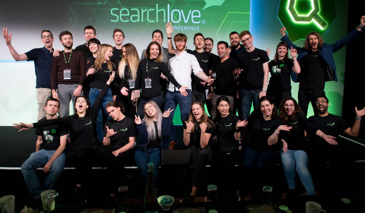 A lively group of people on a stage at the "SearchLove Conference," posing with excitement and making playful gestures, some wearing branded black t-shirts, against a backdrop with a green hexagonal design and conference branding.