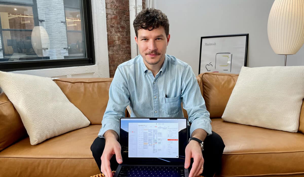 A man with curly hair and a light blue shirt is holding a laptop open to a productivity application, sitting on a tan leather couch in a room with exposed brick and a large window.