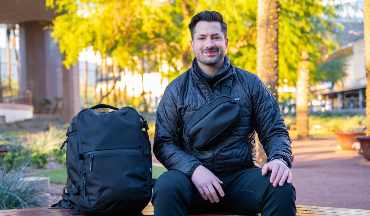A smiling man with dark hair and a beard is seated outdoors on a bench, wearing a black jacket and a black sling bag, with a black backpack beside him.