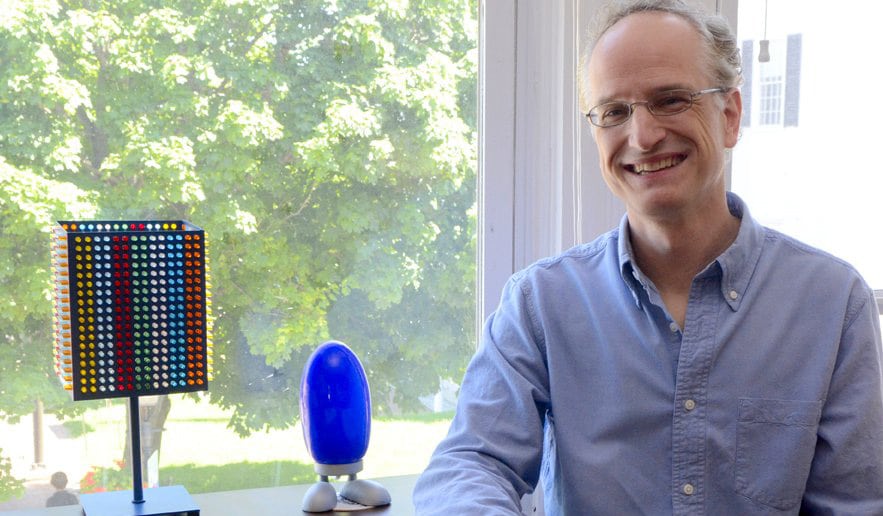 A smiling man with glasses, wearing a light blue shirt, sitting by a window with a view of green trees, beside a desk with colorful, modern lamps.
