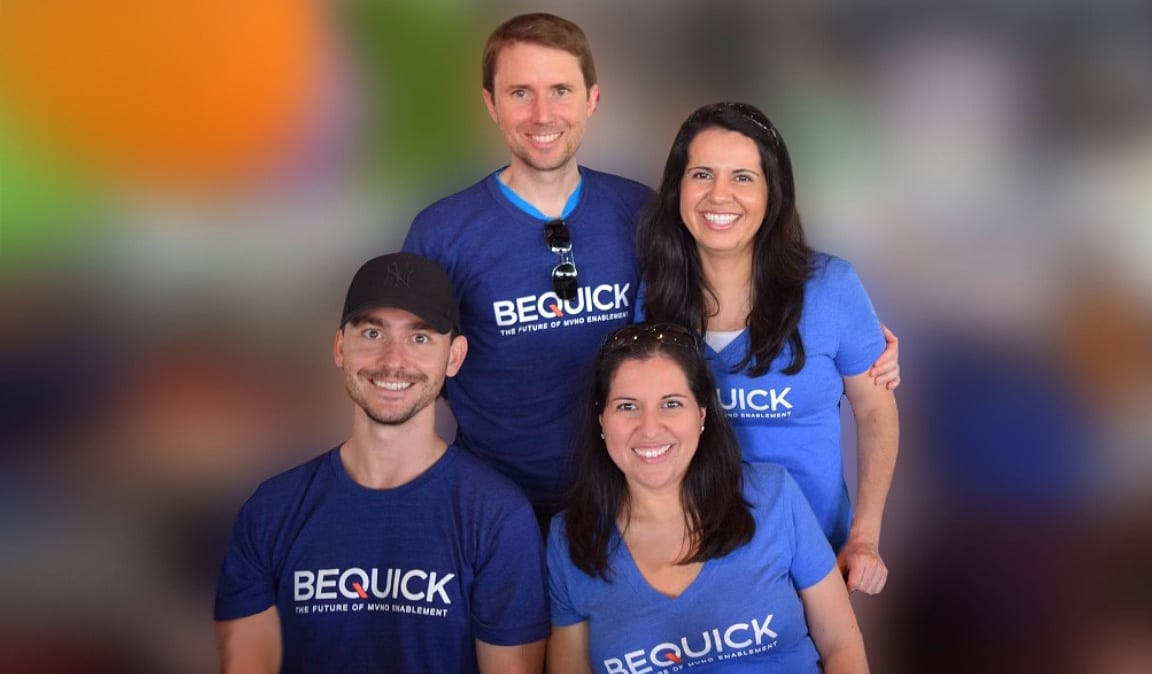 Two couples, each wearing matching blue t-shirts with the logo "BEQUICK," smiling and posing together in front of a blurred colorful background.